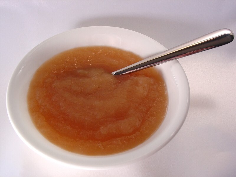 A white bowl of golden applesauce with a silver spoon in it rests on a white surface.