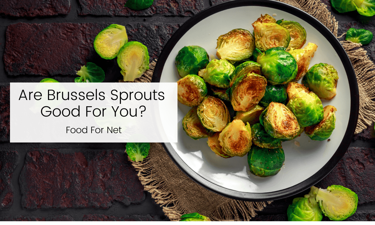 A white plate with grilled Brussels sprouts on a table with a few fresh Brussels sprouts scattered around, looking at whether the vegetables are good for you