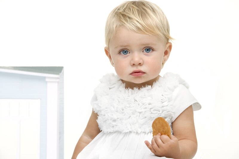 A blonde baby in a white dress, sitting in a white room, holds a cookie in one hand.