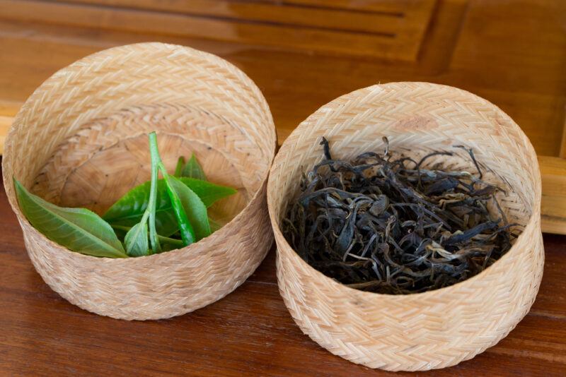 on a wooden surface is a couple of small weaved basket with dried and fresh Asam tea