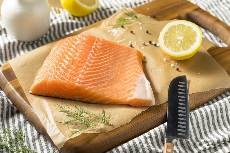 Atlantic-Salmon | Food For Net a slice of fresh Atlantic Salmon on a brown parchment paper with salt, peppercorns, herbs and halves lemons, resting on top of a wooden chopping board with a knife on the side and striped white and grey cloth underneath