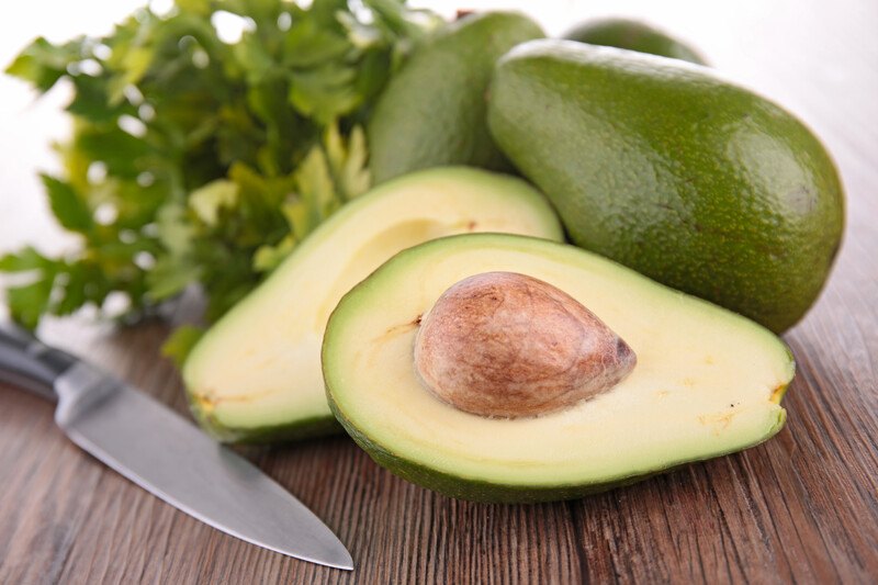 A halved avocado rests next to several whole avocados, a bunch of parsley, and a knife on a wooden surface.