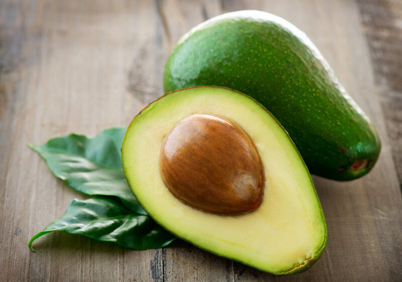 This photo shows an avocado half next to an uncut avocado and some green leaves on a wooden table.