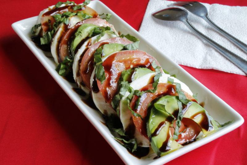 A caprese salad with avocado and balsamic vinegar on a white plate against a red background