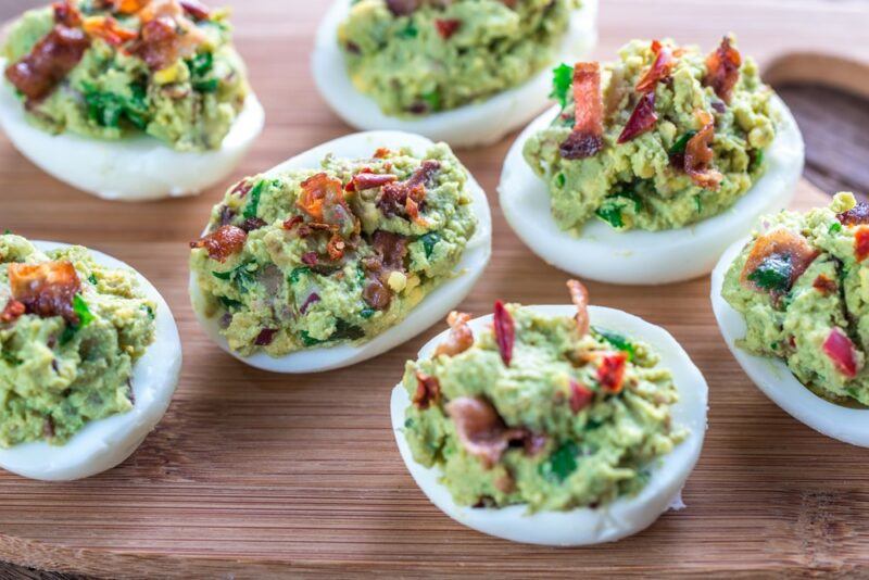 Guacamole deviled eggs topped with bacon on a wooden board