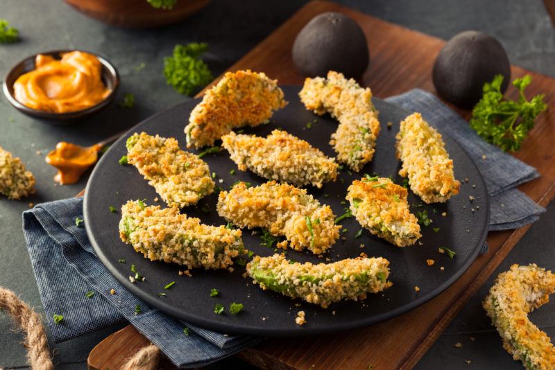 Cooked avocado fries on a black plate next to dipping sauce