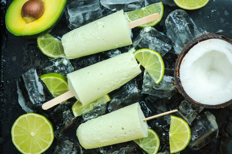 Three avocado popsicles against a black background with ice and lime