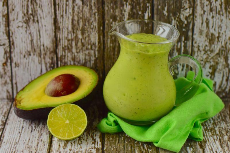 A jug of bright green avocado dressing next to half an avocado and half a lime