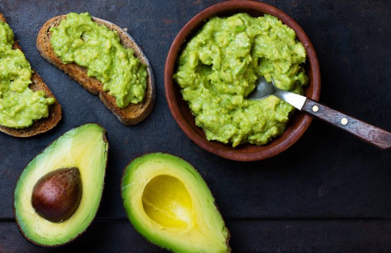 A bowl of mashed avocado, an avocado that has been cut open and avocado on toast