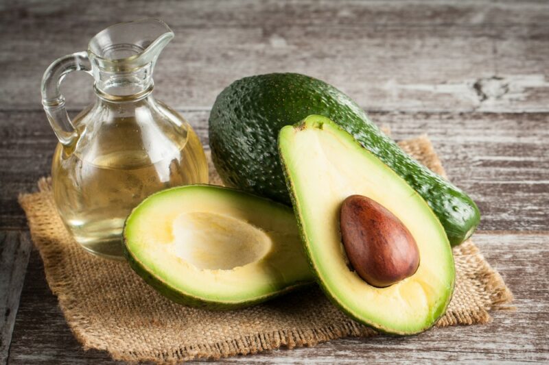 A whole avocado and another that has been cut in half, next to a jug of avocado oil