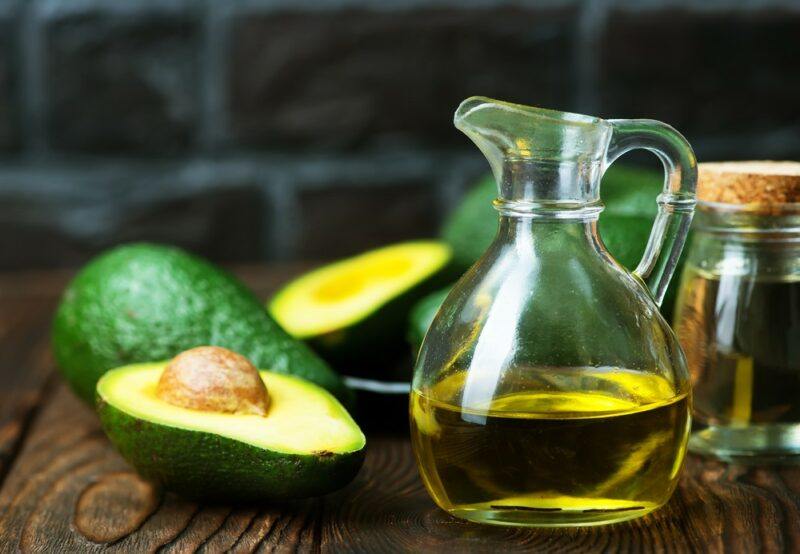 Avocados on a wooden table next to a glass jug with a little avocado oil