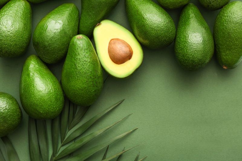 Various whole avocados on a green background with a branch from a tree and one avocado that has been cut in half