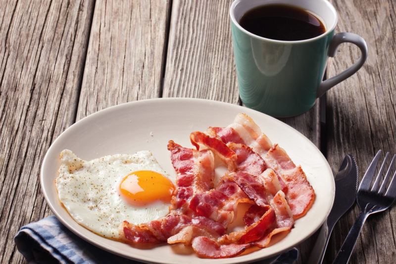 A fried egg and bacon on a plate with a mug of coffee in the background