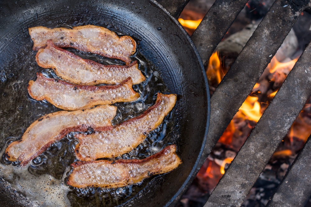 A fry pan with bacon being cooked on a camp fire