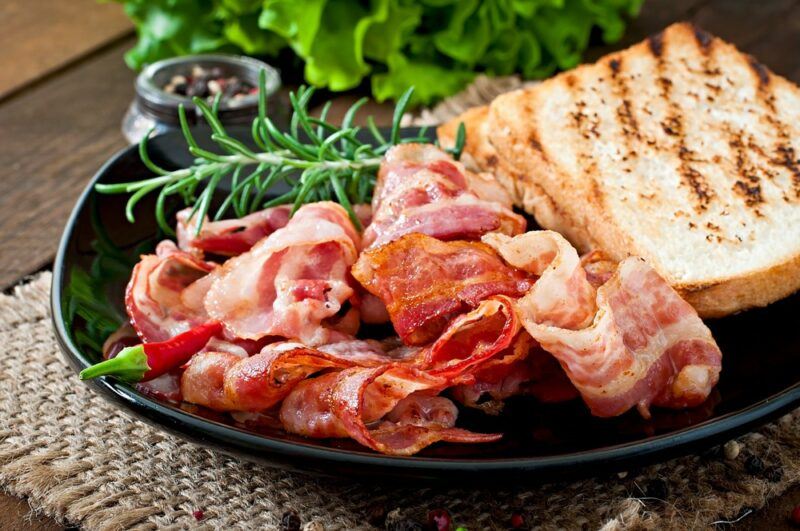 A black plate with cooked bacon, some toast, and some herbs