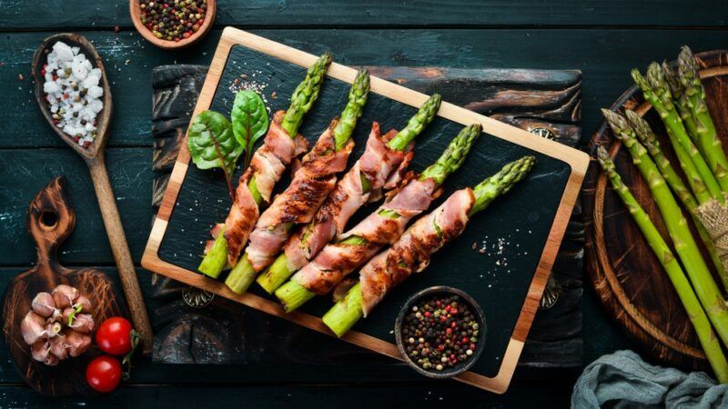 A slate and wood tray with five pieces of bacon wrapped asparagus