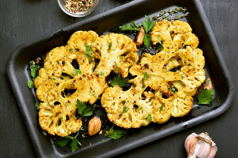 A black baking tray with cauliflower that has been sliced in half, seasoned, and baked