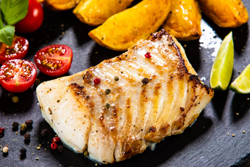 Baked cod on a black background next to tomatoes, potatoes, and lemons