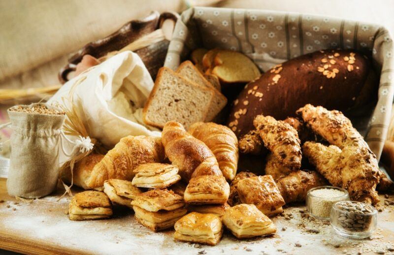 A large basket of baked food that has spilled onto a table. It includes some savory treats and some sweet ones, along with dishes of seeds and a bag of grains