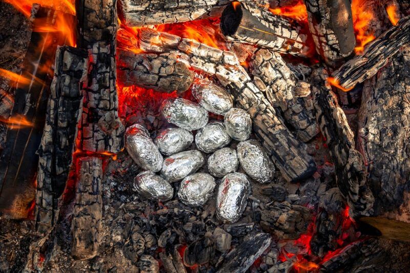 A selection of baked potatoes in tin foil that are cooking in the embers of a fire