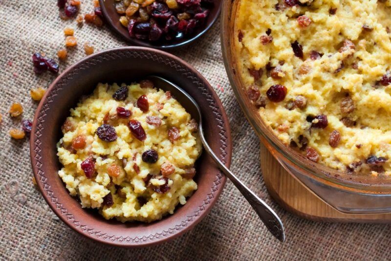 A bowl of baking that uses raisins, next to a larger piece, perhaps bread pudding or raisin bread