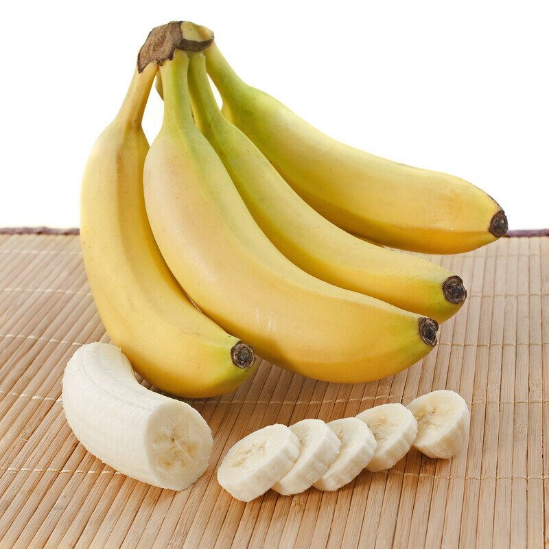 a bunch of bananas and a sliced banana rest on a bamboo mat against a white background.