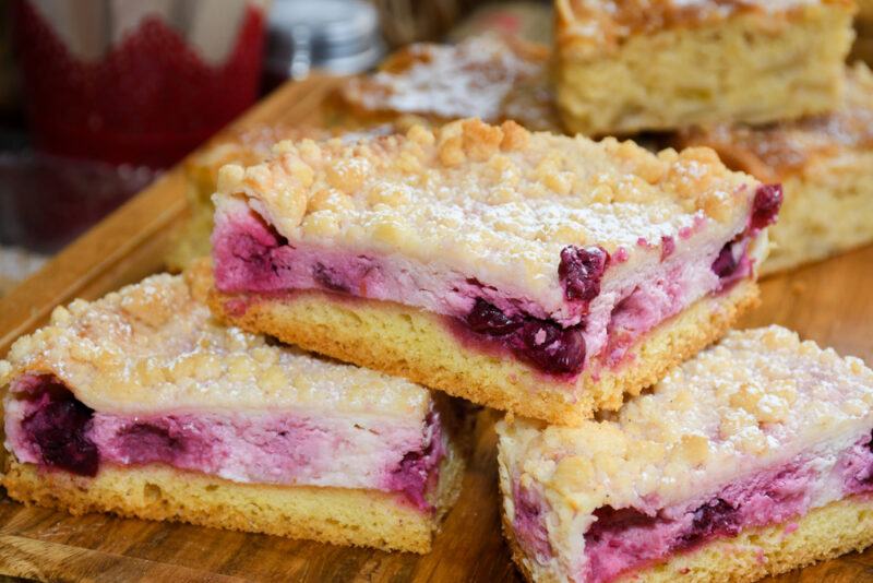 Three cookie bars containing berries and cream cheese rest on a cutting board in front of a stack of a different type of cookie bars.