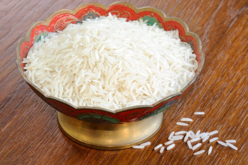 on a wooden surface is an Oriental style bowl with raw Basmati rice, beside it are a few loose Basmati rice grains