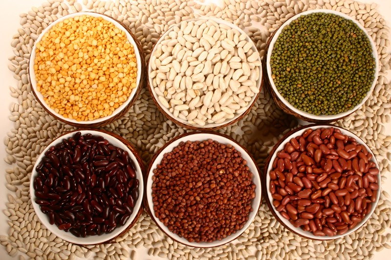 Six bowls containing a variety of legumes in yellow, white, green, brown, and red colors are surrounded by loose white beans.