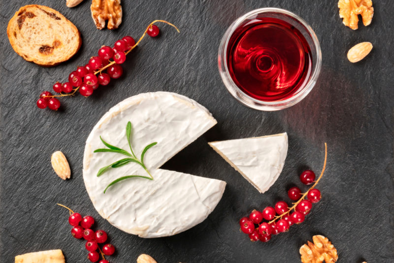 Camembert cheese with a wedge cut out of it, with sprigs of fresh cranberries or currants, bread, and a glass of wine