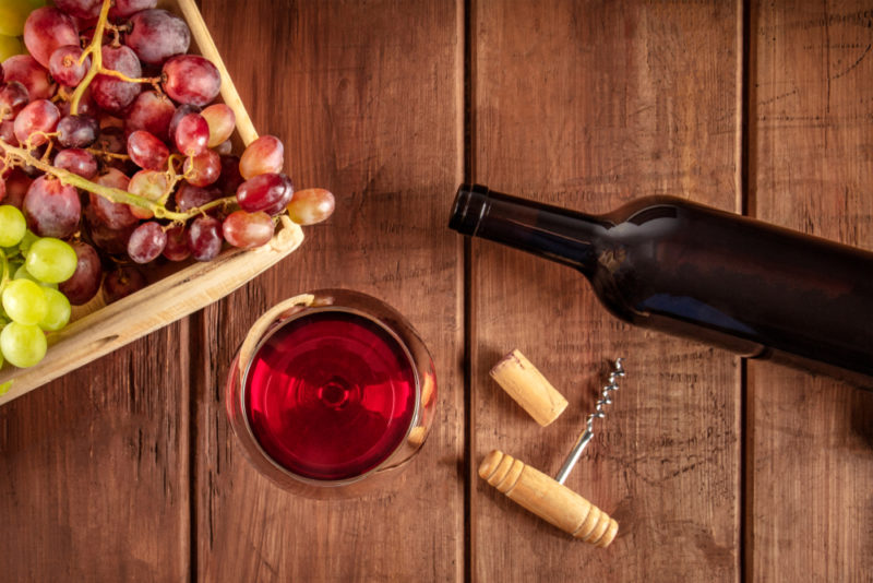 A wooden table with a bottle of Beaujolais wine, a glass of the wine, a cork, corkscrew, and a box of grapes