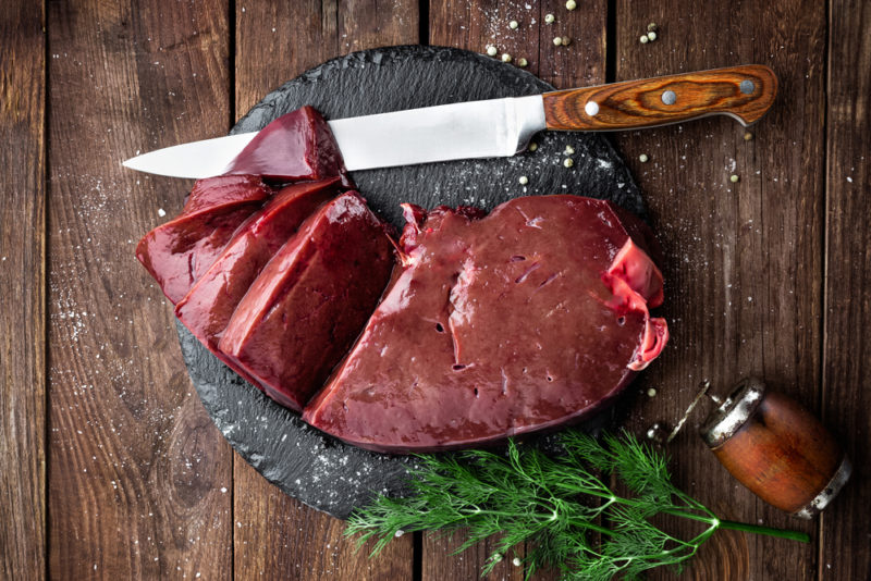 A circular slate with raw beef liver that has been sliced next to a knife on a wooden table