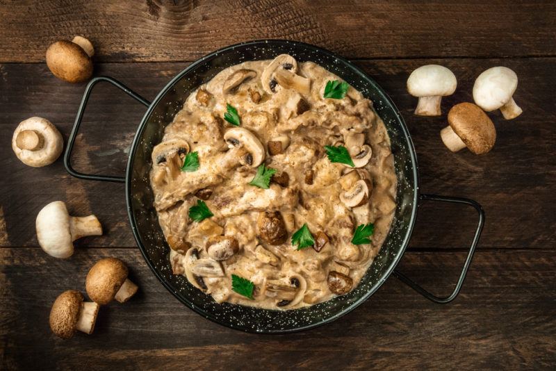 A black bowl with beef stroganoff on a wooden table with mushrooms scattered around