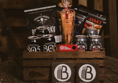 A dark wooden crate containing 6 cans of beer, popcorn and plenty of jerky. There are two coasters resting on the front of it and a bottle opener on top. 