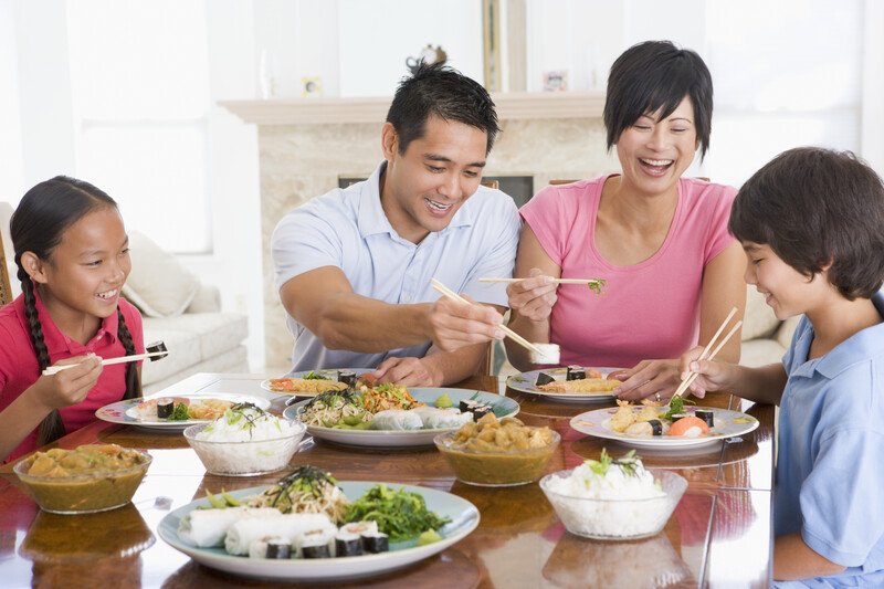 An Asian family with a mother, father, son, and daughter smile as they try seafood, vegetables, and rice dishes at a dining room table, representing the 15 best foods for A Positive blood type.