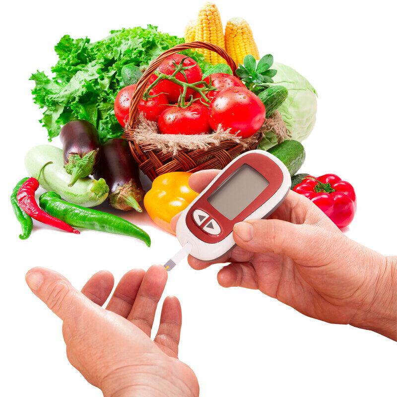 A basket of tomatoes surrounded by several other vegetables rests against a white background behind a set of hands using a device to check blood sugar levels, representing the 12 best foods for lowering blood sugar.