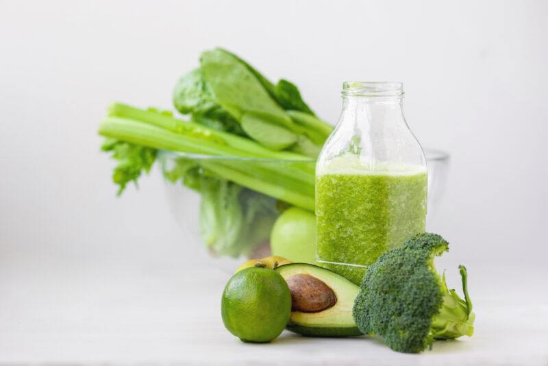 Several green foods, including an avocado, lime, broccoli, and celery, along with a glass bottle filled with a green drink, rest on a white surface with a white background, representing the best foods for detoxifying the body.