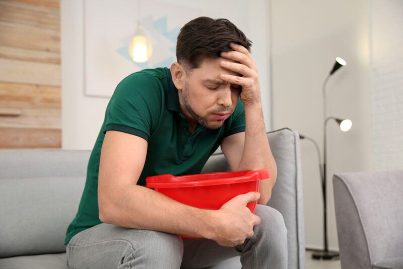 A dark haired, nauseated man in gray pants and a green shirt sits on a gray couch with a red bowl, representing the best foods to eat for nausea.