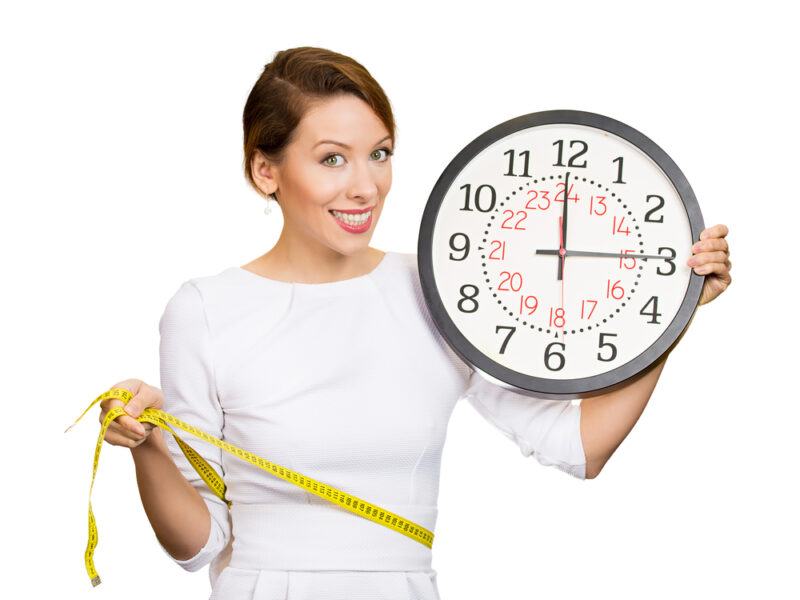 This photo shows a smiling brunette woman in a white shirt holding a yellow tape measure and a clock, representing the best foods for fast weight loss.