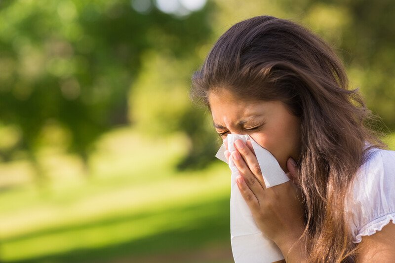 This photo shows a beautiful young brunette woman in a white shirt sneezing into a white tissue in a park, representing the best foods for allergies.