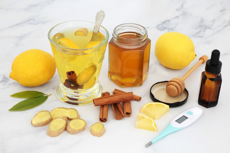 This photo shows two lemons, some cinnamon sticks, a thermometer, two bay leaves, some ginger slices, a jar of honey, a dark brown dropper bottle, and a glass filled with a yellow liquid containing lemon slices, cinnamon sticks, and a spoon, representing the best foods for chest congestion.