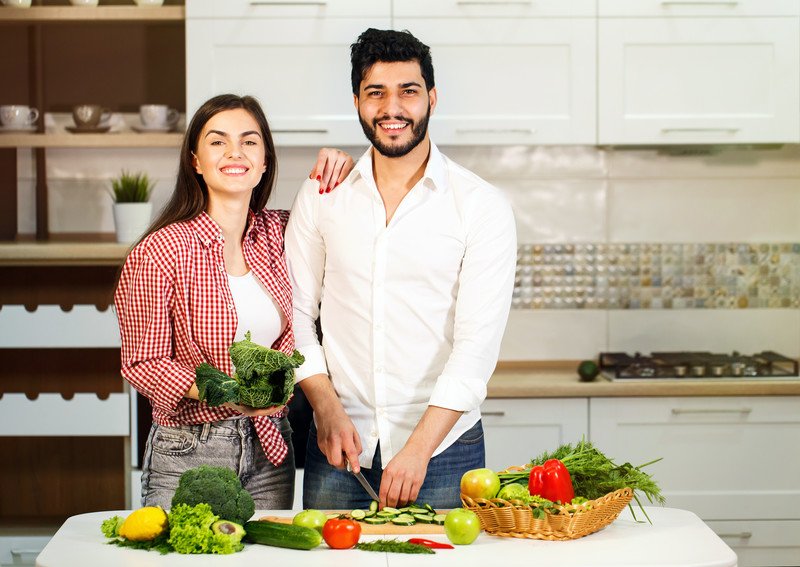 Best Foods For Meal Prep | Food For Net This photo shows a smiling brunette woman in a white and red checked shirt over a white tee shirt and a smiling dark-haired bearded man in a white shirt, standing in a kitchen beside an island filled with broccoli, a tomato, lettuce, an avocado, and various other veggie and fruits on the white table and in a basket, while the man slices a cucumber on a cutting board in front of him, representing the best foods for meal prep.