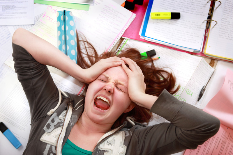 This photo shows a young stressed woman lying on top of a pile of homework, screaming with her eye shut and her hands on her forehead, representing the best foods for stress.