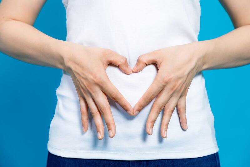 This photo shows the midsection of a woman in a white shirt making an outline of a heart on her stomach with her fingers, representing the best foods for gut health.