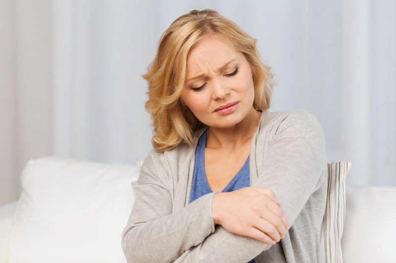 A blonde woman in a blue shirt and gray sweater grimaces as she holds her elbow, representing the best foods for joint pain.