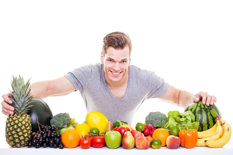 Best Foods Men's Health | Food For Net This photo shows a smiling young man in a gray shirt behind a row of several fruits and vegetables, representing the best foods for men's health.