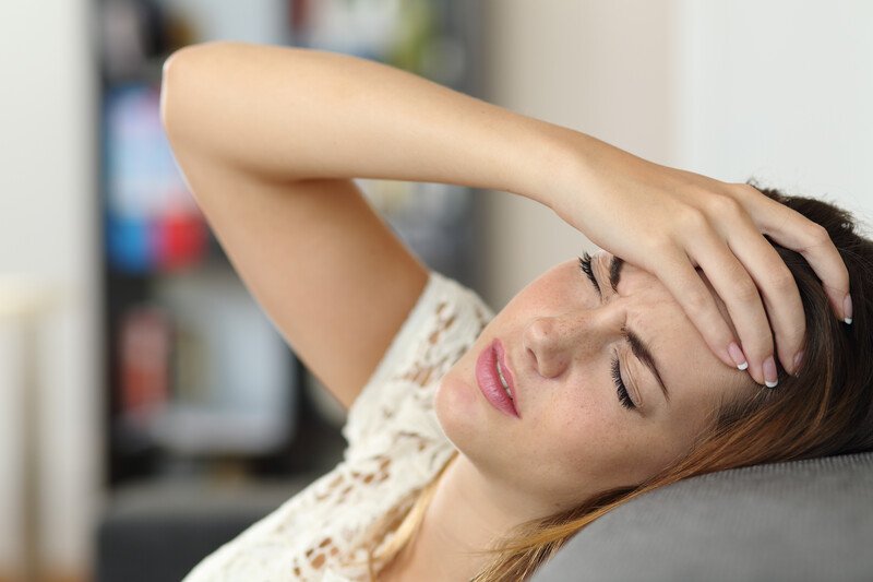 Best Foods Migraines | Food For Net A woman in pain lies back against a gray couch with her hand to her forehead, representing the best foods for migraines.