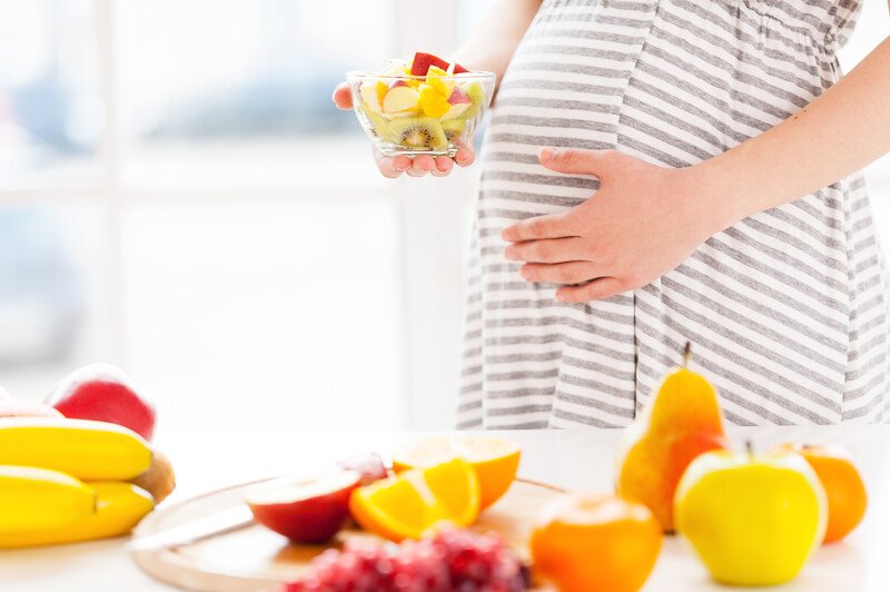 A pregnant woman in a gray and white striped shirt holds a transparent bowl of fruit in one hand while she rests her other hand on her belly, behind a white table with a pear, apples, bananas, and other fruit resting on it, representing the 12 best foods to eat for morning sickness.