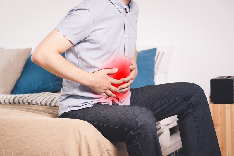 This photo shows a neck-down view of a man in jeans and a gray shirt sitting on a bed, holding his abdomen, which is red as if in pain, representing the best foods for pancreatic diet success.