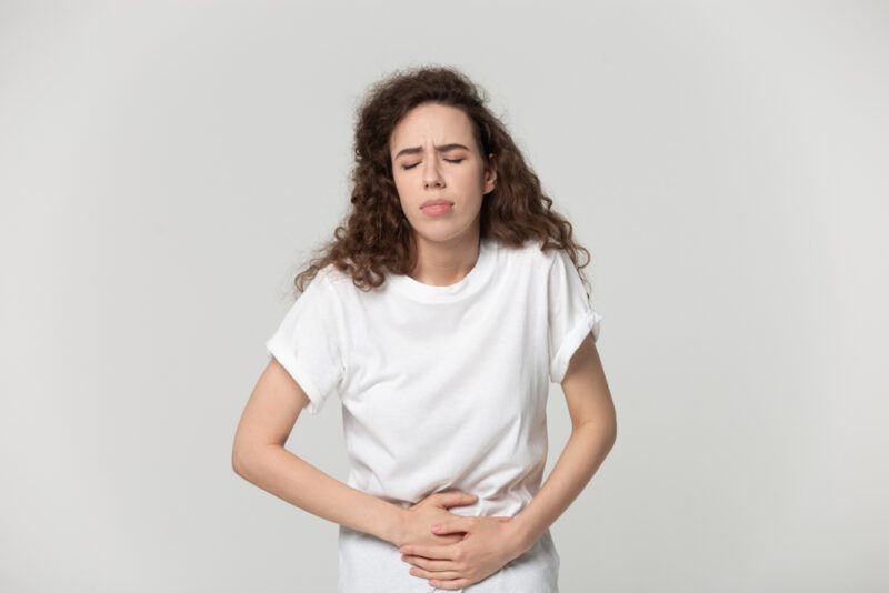 Frown,Girl,Touch,Stomach,Suffers,From,Severe,Ache,Abdominal,Pain | Food For Net This photo shows a brunette woman in a white shirt, grimacing and holding her stomach, representing the best foods for sour stomach issues.